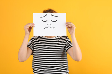 Woman hiding behind sheet of paper with sad face on yellow background