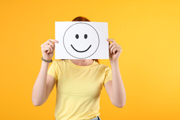 Woman hiding behind sheet of paper with happy face on yellow background