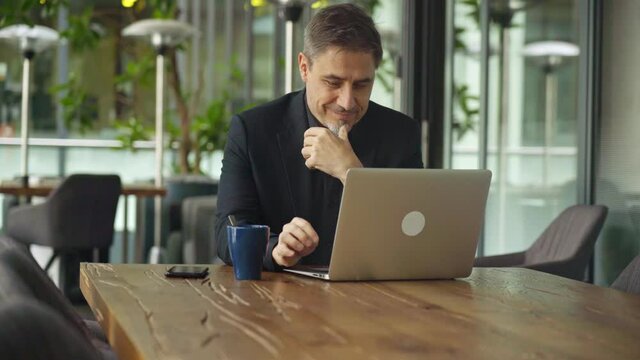 Business portrait - businessman sitting in in office working with laptop computer, thinking. Mature age, middle age, mid adult man in 50s with happy confident smile. Copy space.