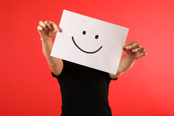 Woman hiding behind sheet of paper with happy face on red background