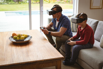 Caucasian father and son wearing vr headsets playing video games sitting on the couch at home