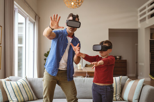Caucasian father and son wearing vr headsets gesturing while standing in the living room at home