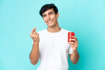 Young Argentinian man holding a refreshment isolated on blue background making money gesture
