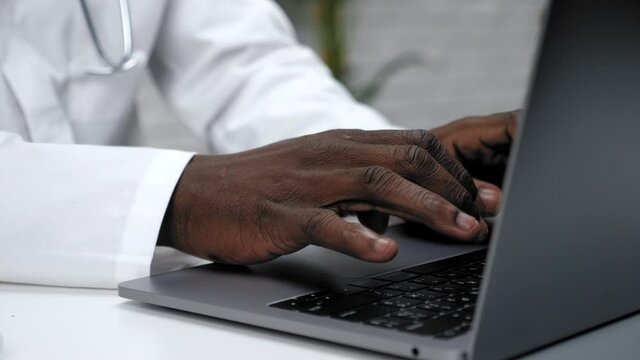 Close up African American hands typing text on the keyboard laptop in clinic. Man doctor in medical gown sitting on chair at desk working and uses computer at medical hospital office