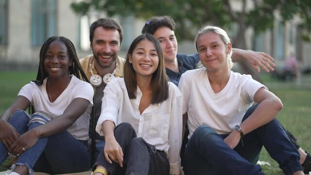 Friendly group of cheerful multinational college students waving hello while sitting on campus lawn. Joyful multiracial classmates smiling while posing for joint portrait video after studies