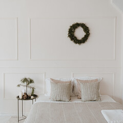 Modern bright home bedroom interior with bed, neutral bed linens, pillows, Christmas wreath made of pine needles hanging on white wall. Aesthetic living room with Christmas decorations