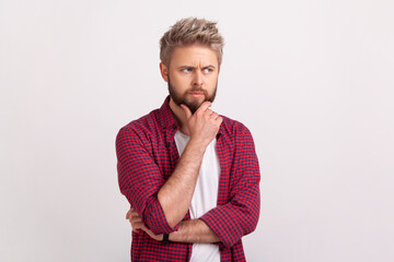 Portrait of young pensive man with beard touching chin and thinking about important question, making hard decision. Indoor studio shot isolated on gray background