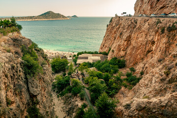 View of an azure sea lagoon with a beach and green trees at the bottom of a deep canyon. Car track on the side of a cliff. Mountain island in the background