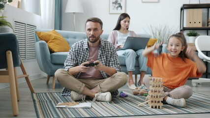 Father and little daughter are playing video game while mother is using laptop indoors at home. Leisure time activities and family life concept. - Powered by Adobe
