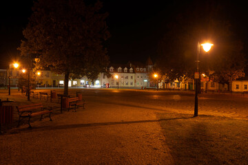 Centralny plac w małym miasteczku nocą, oświetlony światłem latarni ulicznych.