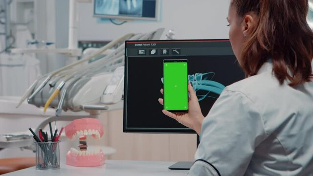 Dentist vertically holding smartphone with green screen while looking at teeth scan on computer in office. Teethcare specialist using mobile phone with chroma key for isolated background