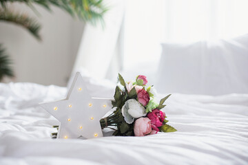 Glowing LED star and a bouquet of artificial peonies on a white bed in the interior. - 463839990