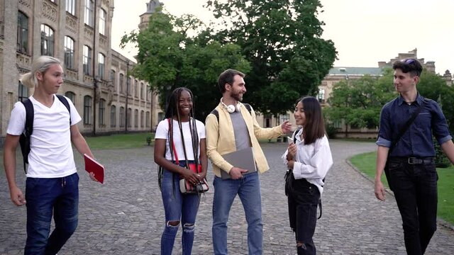 Joyful bearded guy chatting with asian and african american girls in campus park when two classmates coming up they all greeting their elbows. Diverse students enjoying free time before study outdoor