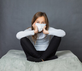 Portrait of a young teen girl in a medical mask sitting on the ground with grey background. - 463842779