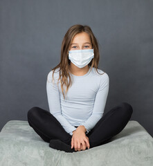 Portrait of a young teen girl in a medical mask sitting on the ground with grey background. - 463842783