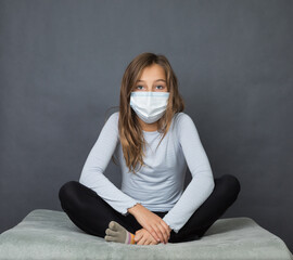 Portrait of a young teen girl in a medical mask sitting on the ground with grey background. - 463842786
