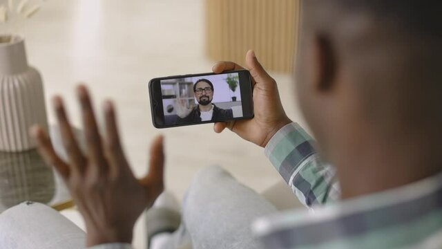 Back view of young African American man waving hello in webcam of smartphone, while having video call with his Caucasian male friend at home indoors. Meeting online, remote work from home