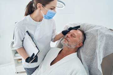 Beautician in medical mask examining man forehead