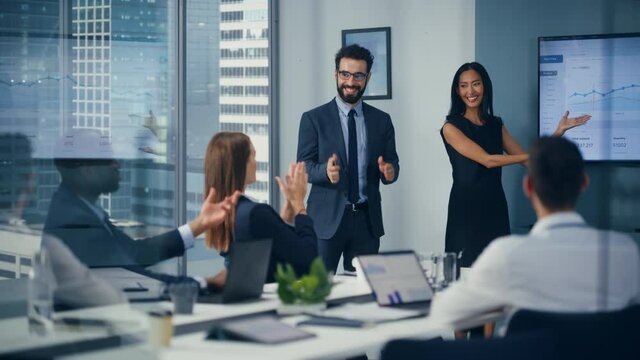 Diverse Office Conference Room Meeting: Male Executive and Asian Female Top Manager Present e-Commerce Startup Growth Statistics on Wall TV to a Group of Investors. Everybody Celebrates Great Results