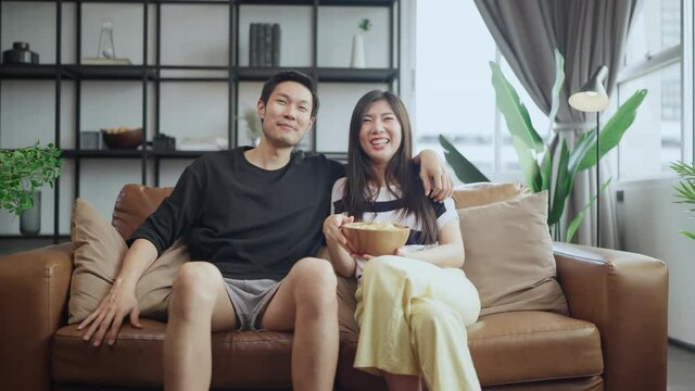 Asian sweet couple watching tv relax sitting on sofa in living room at home. wife is holding popcorn bowl looking to TV.asian couple stay home,asian family Relationship enjoy movie at home concept.