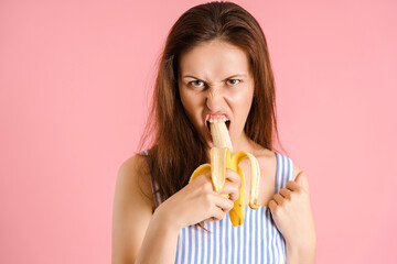 A very hungry sexy brunette aggressively bites a banana on a pink background