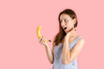 A confused woman holds a banana with a condom on it. The woman opened her mouth in surprise while standing on a pink background, copyspace