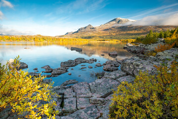 Mountain and sky reflecting in lake in early morning of autumn. Autumn colors in remote arctic landscape. Nieras mountain reflects in Lulealven dam in Stora Sjofallet national park, Sweden.