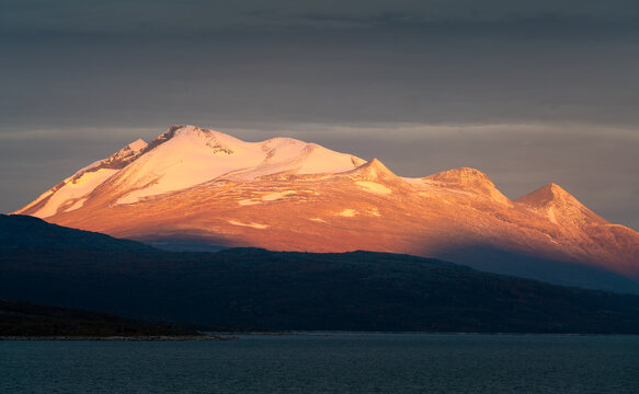 Epic sunrise light illuminating rocky, glacier covered peak with dark cloudy sky in the background. Ahkka peak in beautiful golden hour light, Stora Sjofallet National Park, Sweden. Arctic landscape.
