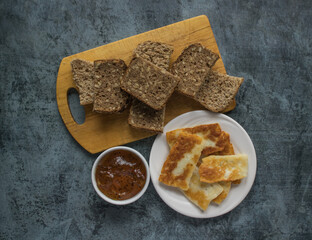 Fried halloumi, haloumi, hellim cheese on a white plate next to sliced homemade bread with seeds and fruit jam on a dark background. Traditional food. Cypriot cuisine