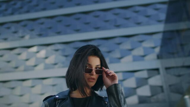 Confident young brunette woman in trendy stylish look wearing black leather jacket and sunglasses looking at camera standing by building in city street, touches her hair. Gimbal shot, steadicam