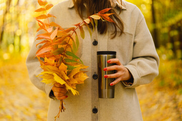 A woman is holding a thermo cup and a bouquet of autumn leaves. Autumn outdoor activity. Autumn day