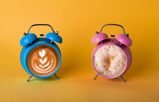 Two alarm clocks with cappuccino and latte inside on yellow background. Time to drink coffee concept. Choice between two type of coffee