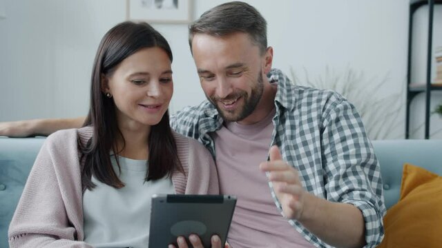 Cheerful young couple husband and wife are using tablet smiling touching screen and talking indoors at home. Technology and people concept.