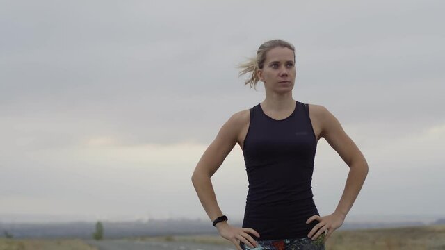 Woman stands with confident look, looks at smartwatch on hand, starts run background overcast sky. Woman is jogging outdoors