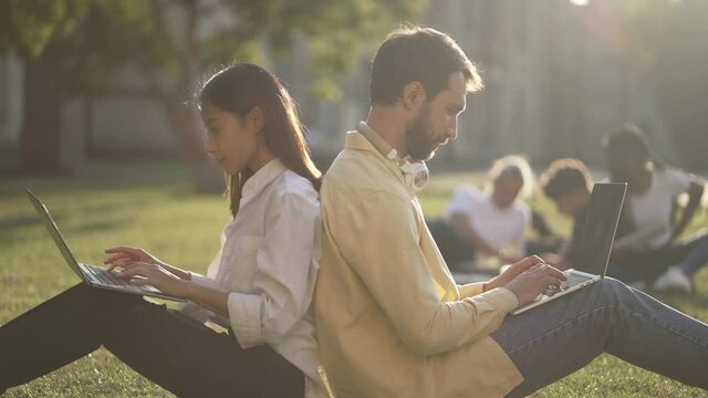 Two multiracial classmates, young asian female and caucasian male, networking on laptops while sitting back to back on sunlit campus lawn. Multi-ethnic students preparing for exam outdoors