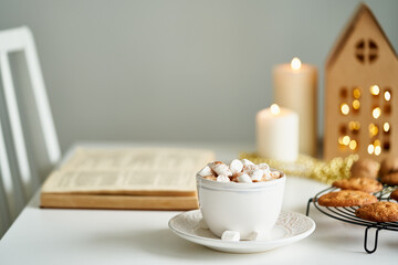 Christmas light gray background with copy space. Reading book on cozy winter evening with candles, cocoa hot chocolate on grid with marshmallows and cookies. Side view of table, chair in home room. 