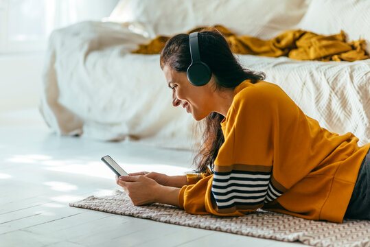 Happy woman in casual yellow clothing lying down floor and using phone for video call at her home. Person having leisure time indoor looking at smartphone and browsing the internet.