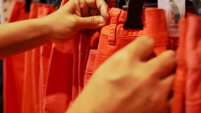 Female hands choosing at the size of orange jeans on hanger in a clothing store.