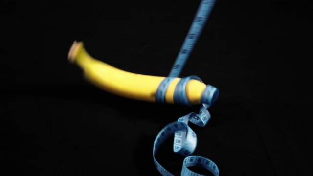 A pair of hands in black leather gloves removing tailor's tape wrapped on a ripe yellow banana-. Visual metaphor: checking the size of a specificl human anatomical part.
