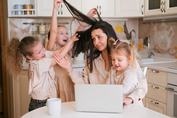 Young mother sitting with laptop on kitchen and three daughters interfere her work