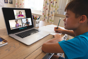 Smiling caucasian boy using laptop for video call, with diverse elementary school pupils on screen
