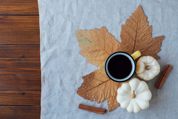 Autumn flat lay with a cup of hot drink, white mini pumpkins, dry maple leaf and cinnamon sticks on neutral tablecloth and wooden rustic background. Copy space.