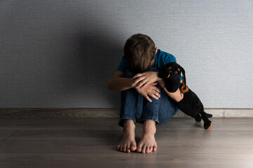 Boy sitting on floor in dark room with face hidden in knees and holding toy dog for feeling of...
