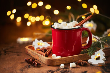 Red cup of hot cocoa with marshmallows on wooden table with christmas tree and glowing garland for...