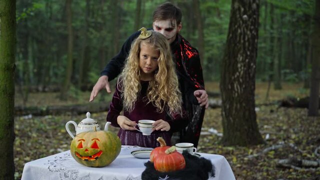 Little girl pouring tea in cup as scary vampire coming from forest at background. Portrait of Caucasian child on Halloween in woods outdoors. Holiday concept