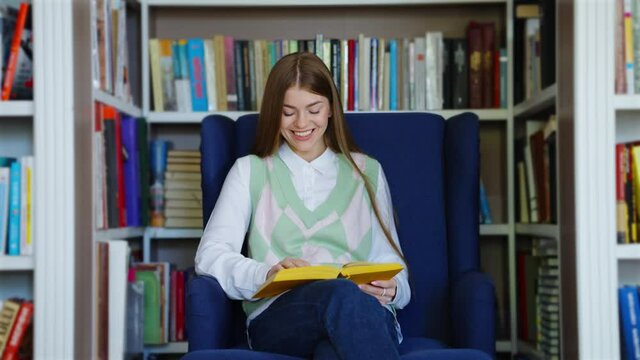 Young female with long hair wearing white shirt and vest sitting in cozy armchair and laughing while reading funny book, bookshelves on background. Reader enjoying comic literature at bookstore