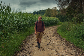 Man with blond hair and a stubble beard walks on a path between a cornfield and a forest in the countryside under a dark cloudy sky.