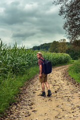 Man with blond hair and a stubble beard walks on a path between a cornfield and a forest in the countryside under a dark cloudy sky. Rear view.