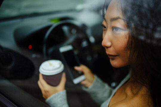Young asian woman drinking coffee while driving her car. Drinking morning coffee while driving to work. Portrait of a young and cheerful woman with coffee cup driving luxury car in the city