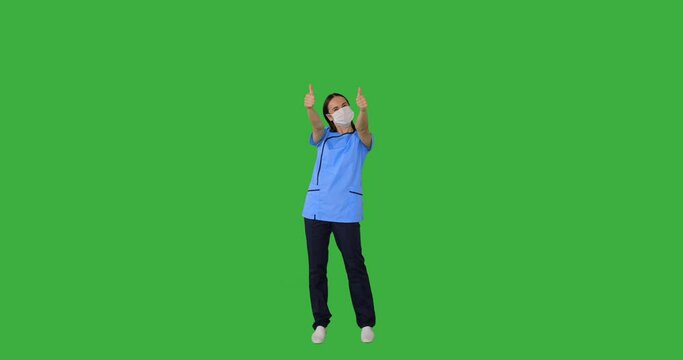 Excited woman nurse wearing protective face mask and giving thumbs up gesture over green background
